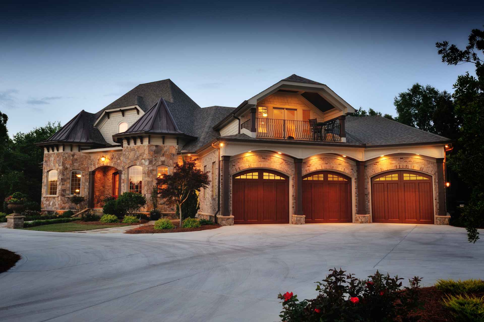Classic castle-style luxury custom home featuring varied multi-tonal stone masonry siding, a dark architectural roofline, warm wooden garage doors, and a spacious paved driveway.