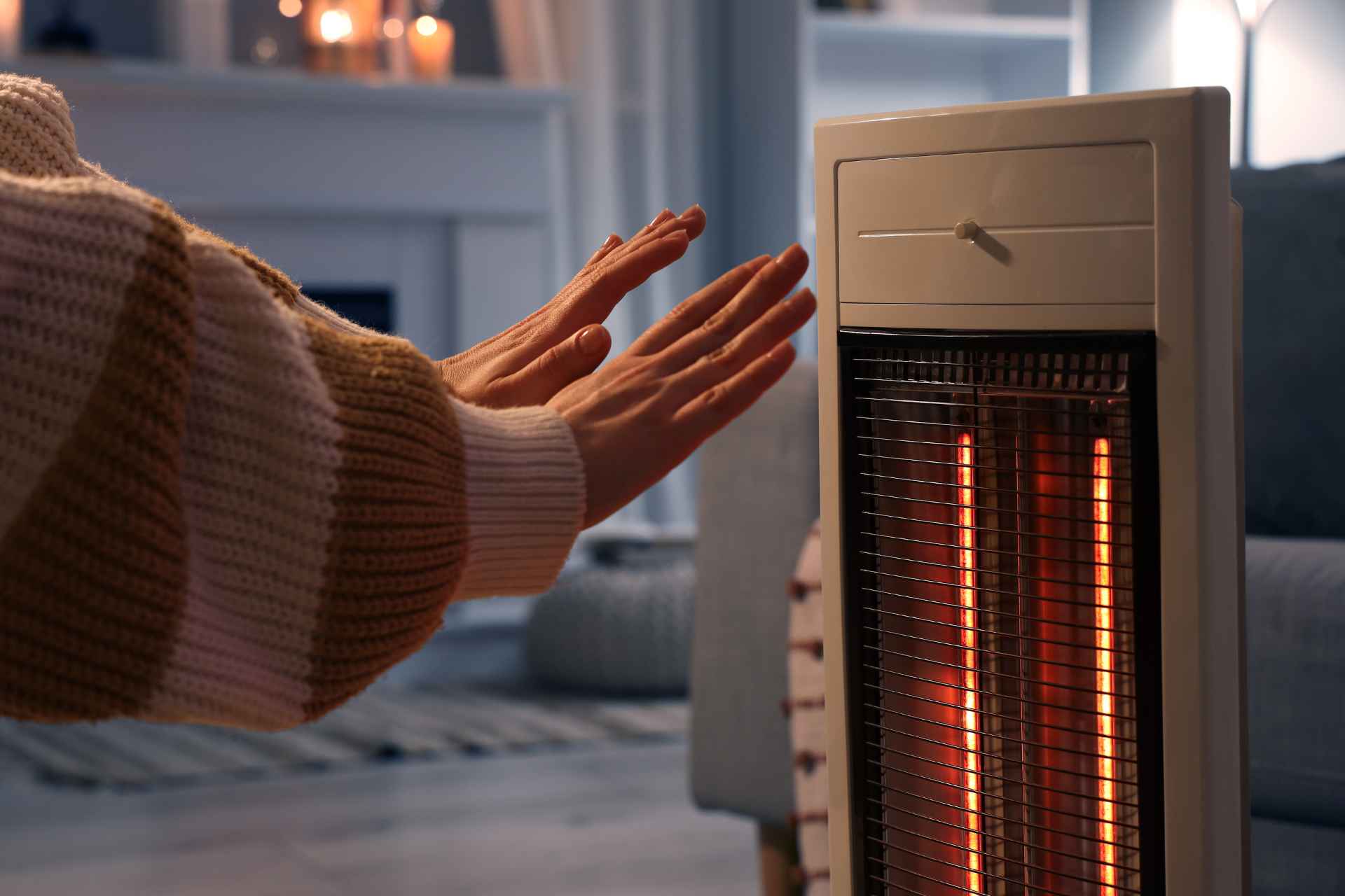 A close-up of a woman warming her hands near an electric heater in a living room at night.